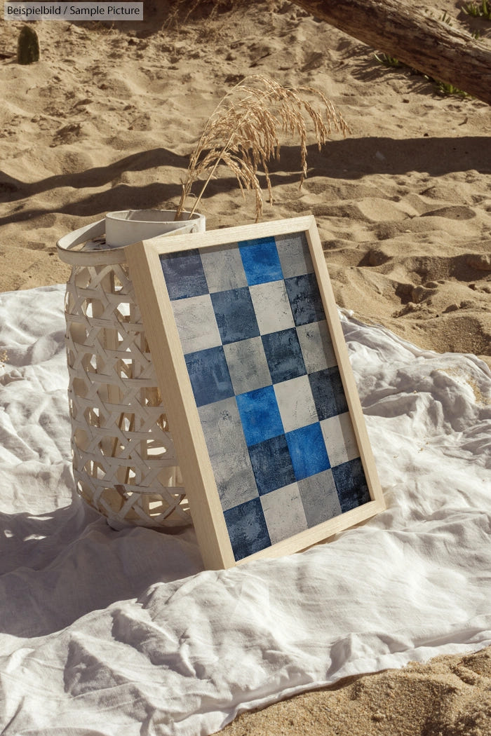 Beach scene with woven lantern, white blanket, and blue checkered art piece. Sunlit sand in the background.