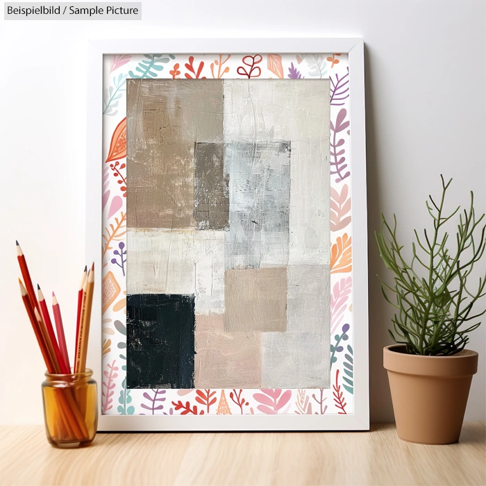 Geometric abstract painting in frame with plant and pencils on patterned background.