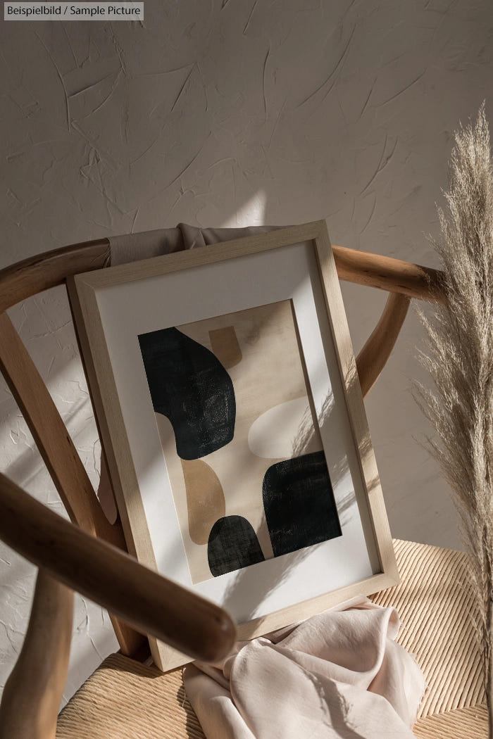 Framed abstract artwork on chair with neutral tones and pampas grass against textured wall.