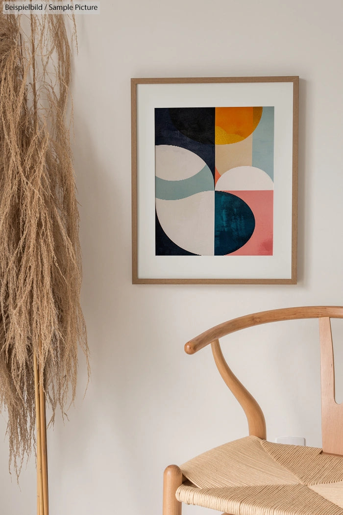 Framed abstract art with geometric shapes and earthy tones on a beige wall, next to a wooden chair and pampas grass.