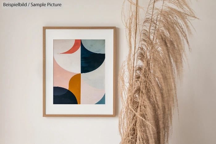 Framed abstract painting with geometric shapes in pink, blue, and yellow beside dried pampas grass against a white wall.