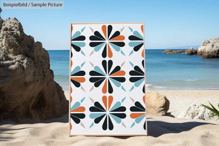 Abstract geometric artwork with orange, black, and blue shapes on a canvas at a sandy beach with rocks and ocean.