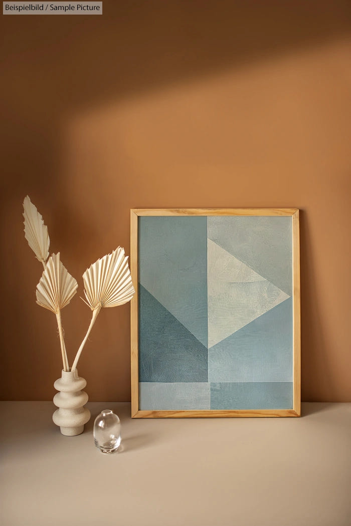 Geometric abstract art in a wooden frame, next to a decorative vase with white leaves, on a warm brown background.