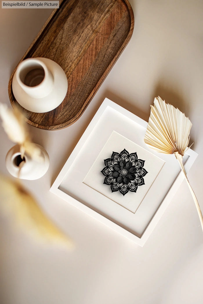 Decorative black mandala art in white frame on table with vases and dried leaves.