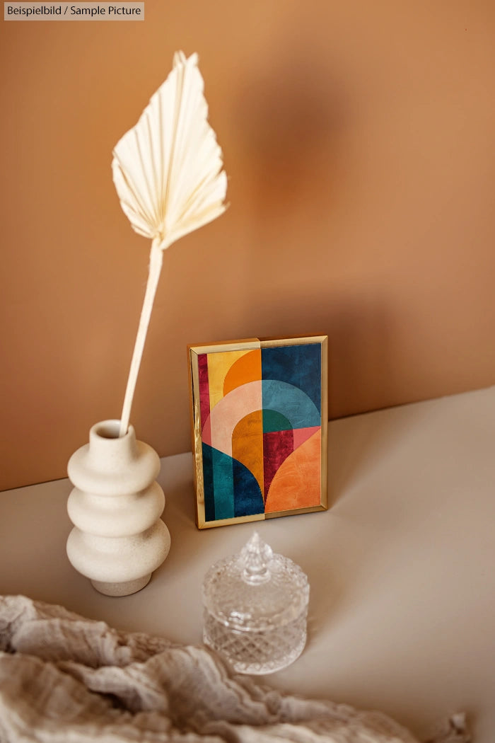 Modern art print with geometric shapes in a gold frame, beside a white vase with a dried leaf, on a beige surface.