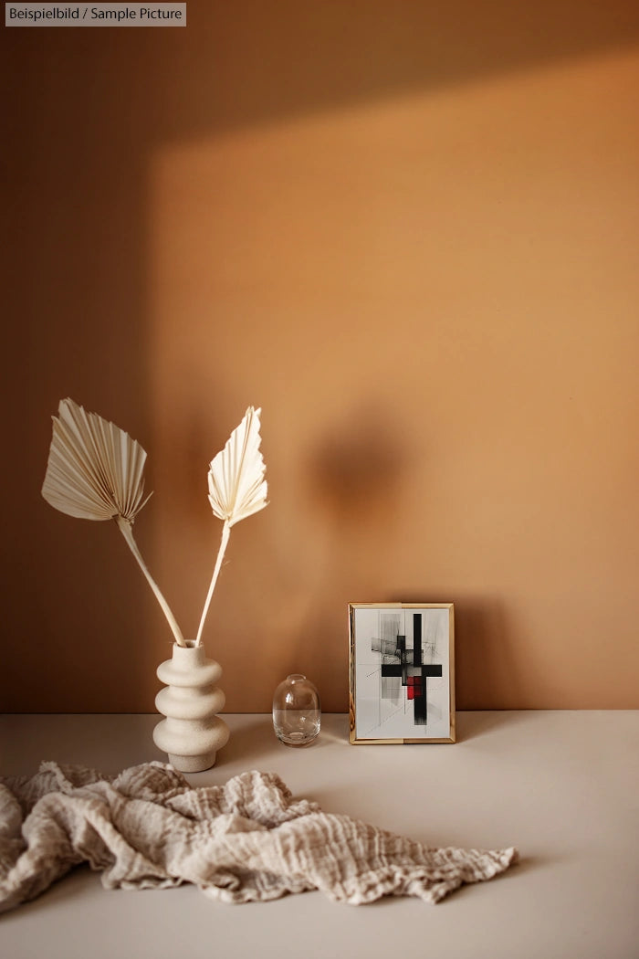 Minimalist beige interior with abstract art, a ceramic vase with dried leaves, a small glass object, and a textured cloth.