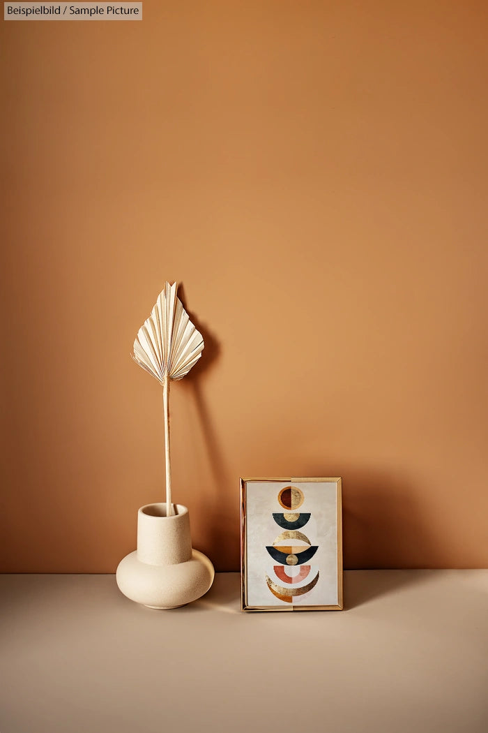 Minimalist decor with a vase and palm leaf, alongside a framed geometric art piece on a beige surface.