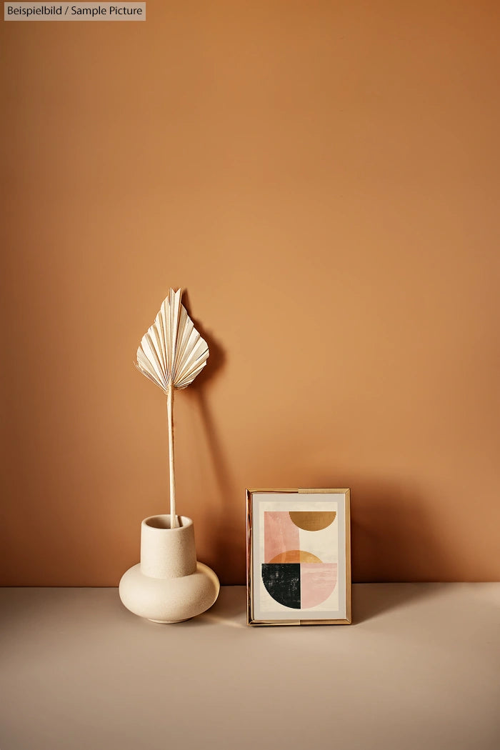 Modern beige vase with dried palm leaf next to framed geometric abstract art on a beige background.