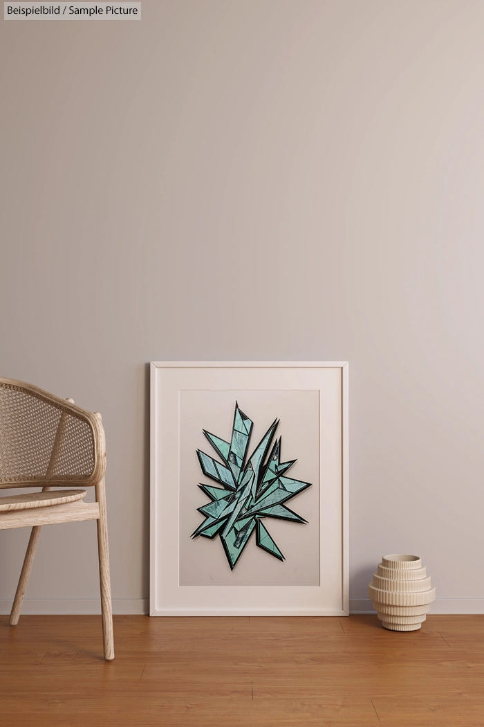 Framed geometric artwork with teal and black shapes leaning against a beige wall, beside a wicker chair and ceramic vase.