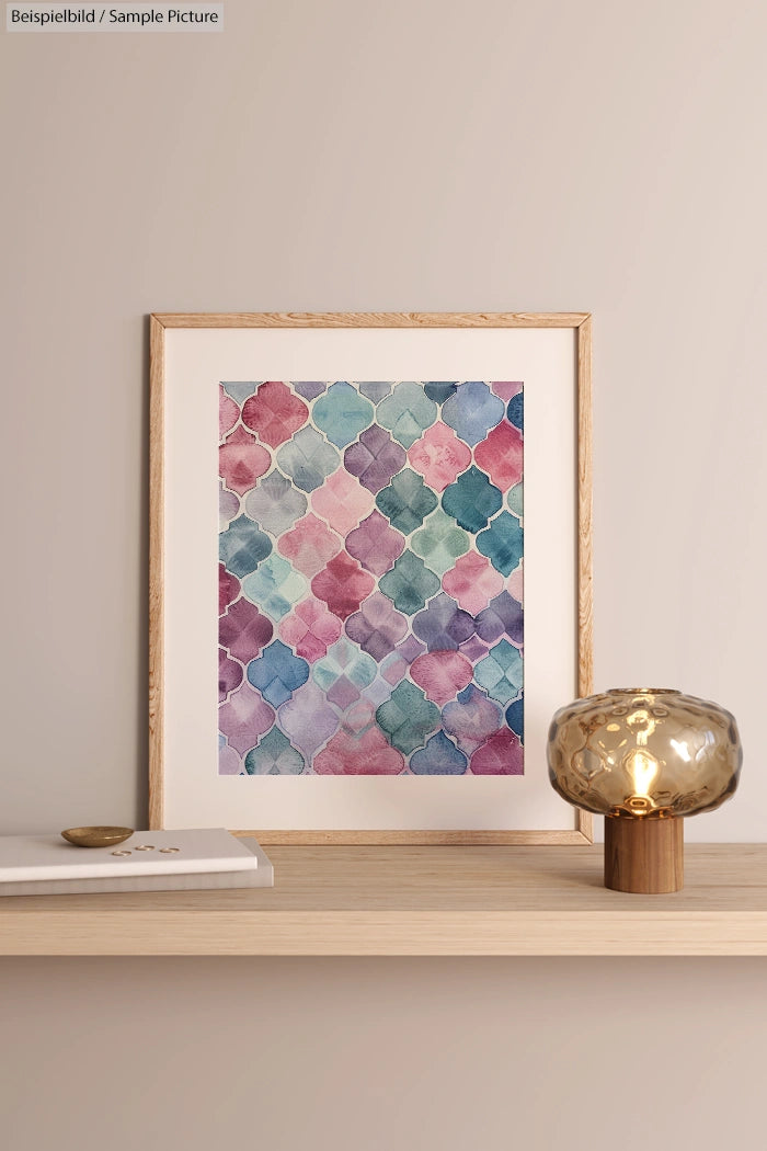 Framed geometric watercolor art in pastel tones on a wooden shelf with decorative glass lamp and book.