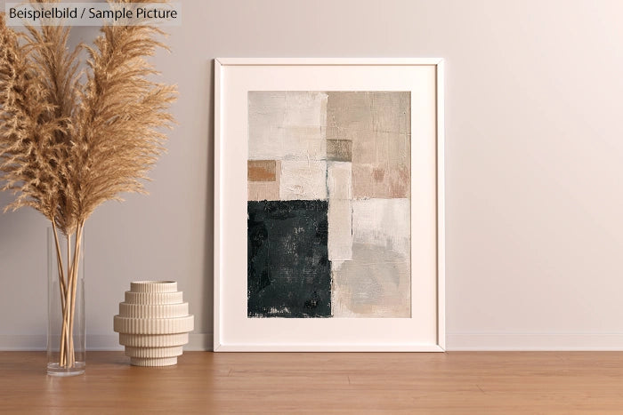 Framed abstract art in beige and black on floor with tall vase and pampas grass against a cream wall.