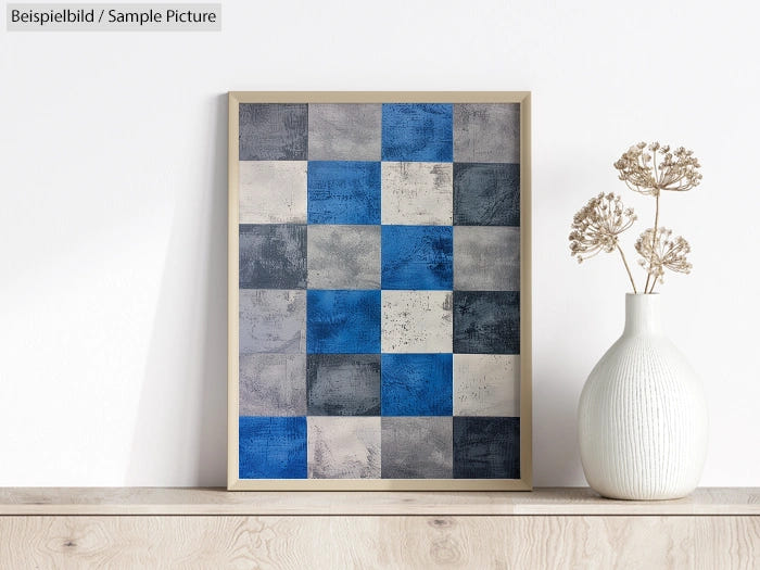 Framed abstract artwork with blue and gray checkerboard pattern, displayed next to a white vase with dried flowers.