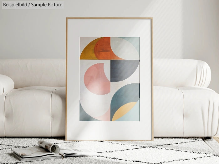 Framed abstract art with geometric shapes in pastel colors leaning against a white couch on a patterned rug.