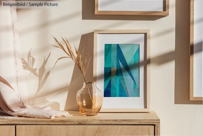 Framed abstract artwork with blue and green shapes on a light wooden cabinet with a glass vase and dried grasses.