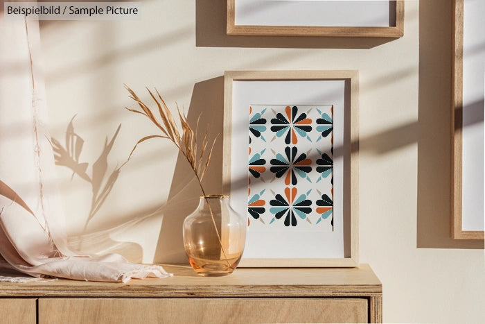 Framed abstract floral art on wooden shelf with dried plants in glass vase, sunlight casting shadows on beige wall.