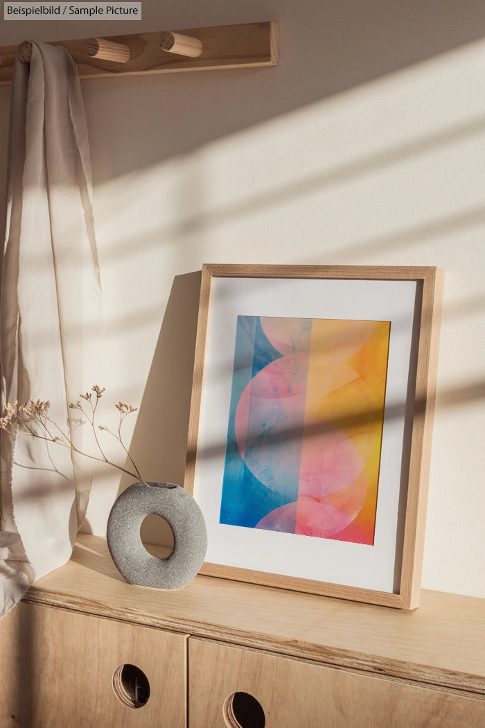 Framed abstract art with pink, blue, and yellow geometric shapes leaning on a wall, next to a vase on a wooden table.