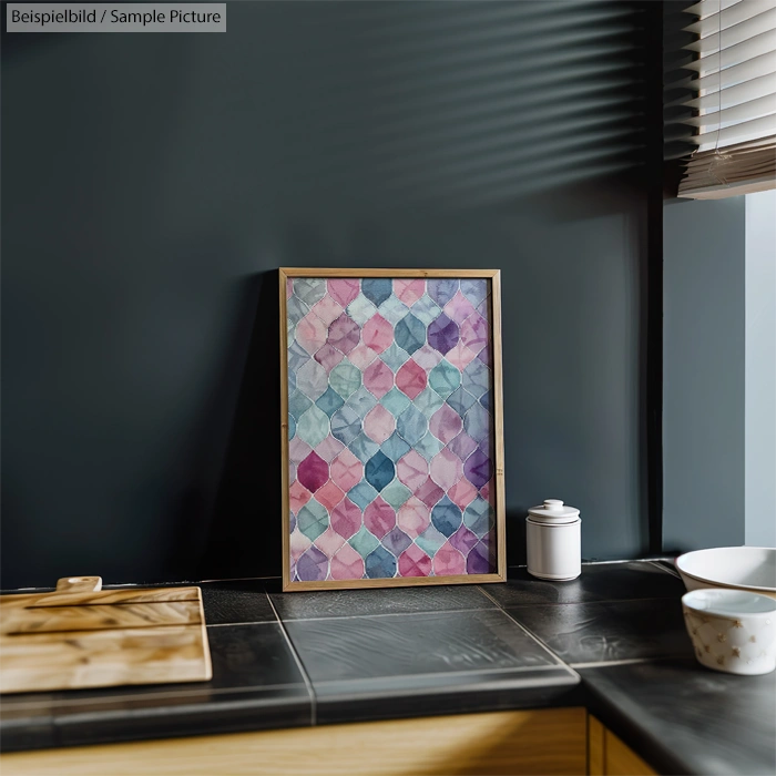 Colorful geometric pattern artwork in wooden frame on dark kitchen counter with cutting board and container.