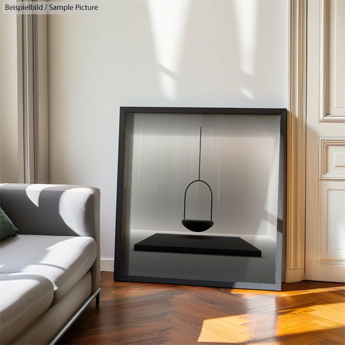 Modern living room with art piece featuring a suspended black bowl in a frame, placed beside a grey couch on wooden floor.