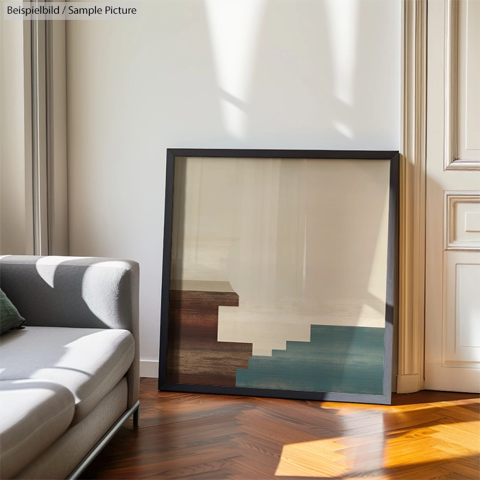 Modern abstract artwork leaning against a wall beside a gray sofa in a sunlit room with wooden flooring.