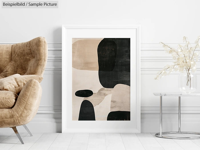 Modern abstract painting with black, beige, and white shapes in a white frame, next to a brown cushioned chair.