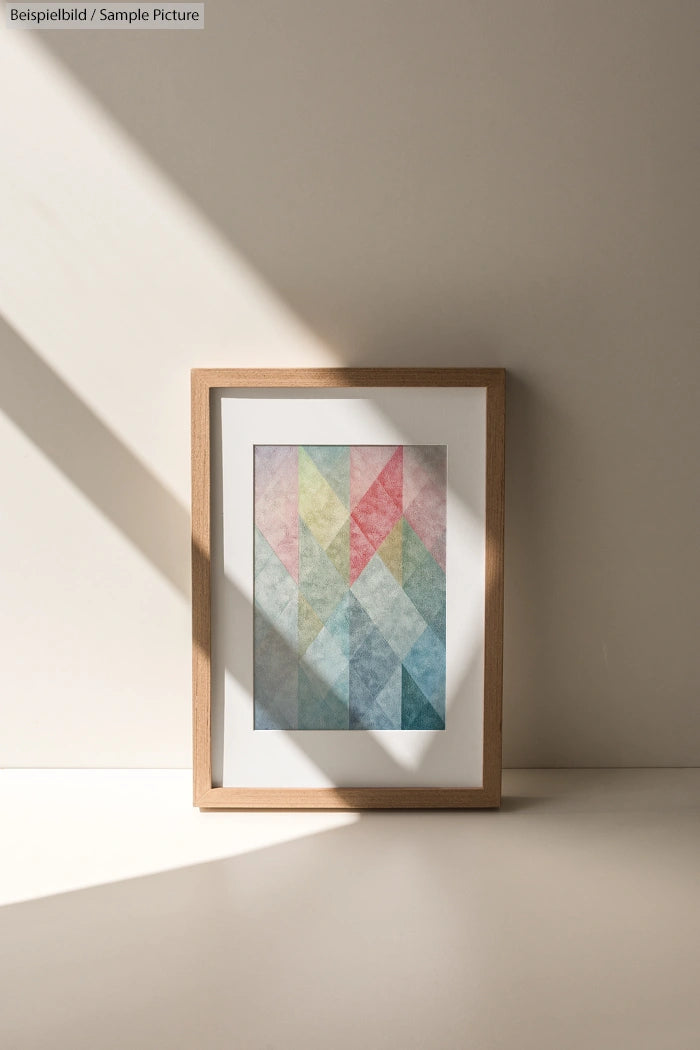 Framed geometric art with pastel colors in a wooden frame, lit by sunlight against a beige wall.