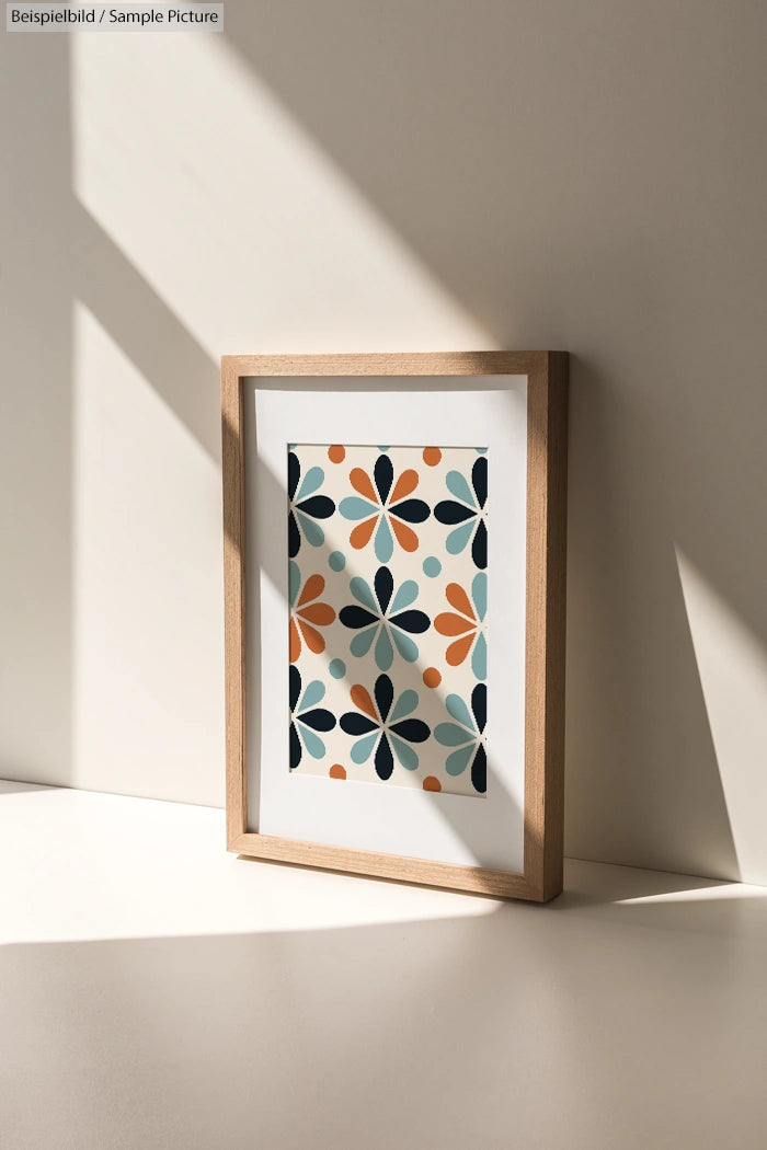 Framed floral abstract art on a sunlit white surface with geometric design in orange, teal, and black.