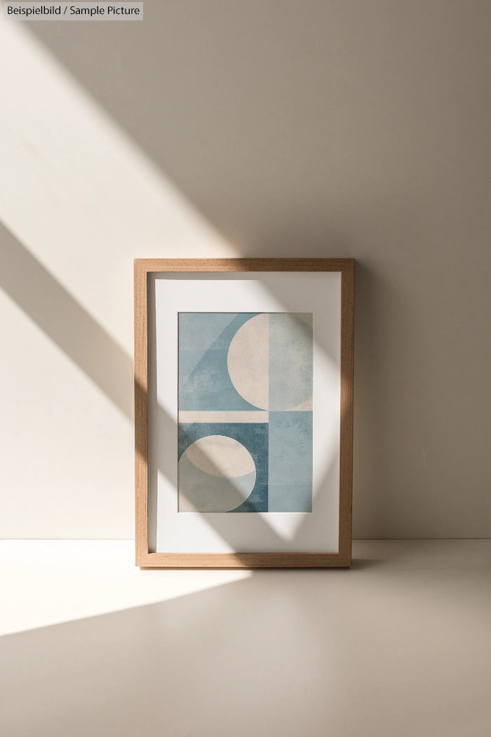 Framed abstract art with circular patterns in soft blue and white, placed against a wall with dramatic sunlight shadows.