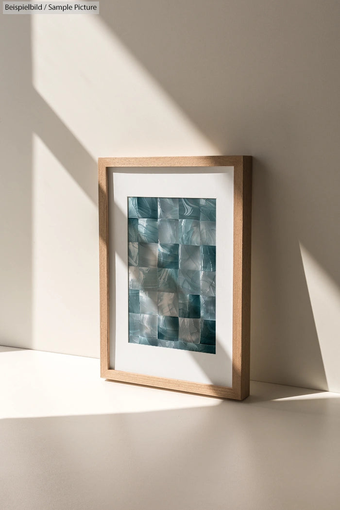 Framed abstract art piece with mosaic-style blue and white tiles on a sunlit table.