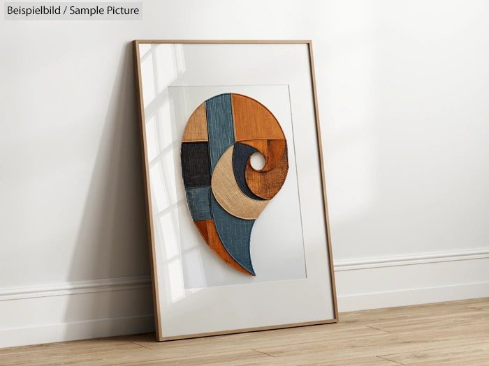 Framed abstract art with a spiral made of wood and fabric pieces, leaning against a white wall on wooden floor.