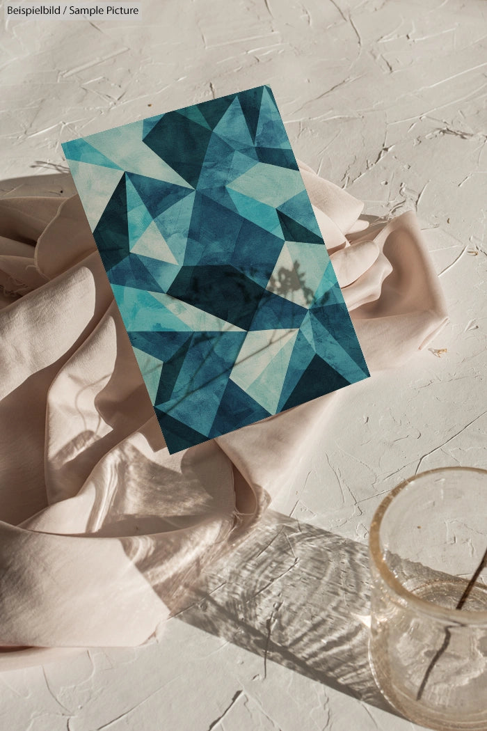 Abstract geometric art in blue and teal tones on textured surface with draped fabric and a clear glass nearby.