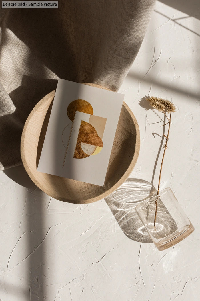 Minimalistic art print in a wooden tray alongside a dried flower in a glass vase, with textured shadows on a white surface.