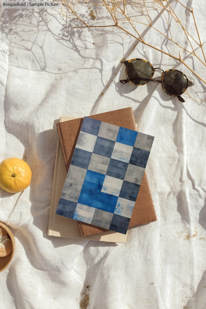 Books on white cloth, topped with a blue plaid notebook, lemon, sunglasses, and twigs in sunlight. Sample image text visible.