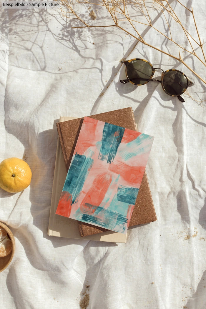 Flat lay of abstract painting on books with sunglasses, lemon, and shadow patterns on white cloth.