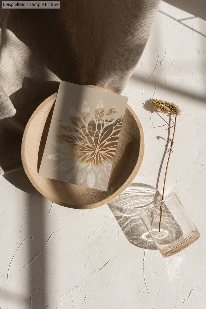 Leaf-patterned card in wooden bowl next to dried flower in vase on sunlit textured surface.