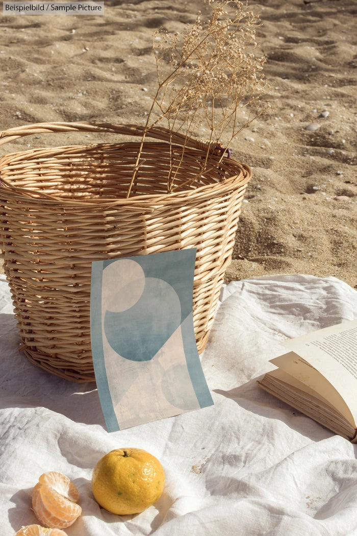 Beach scene with a wicker basket, abstract art, peeled citrus fruit, and an open book on a white cloth.