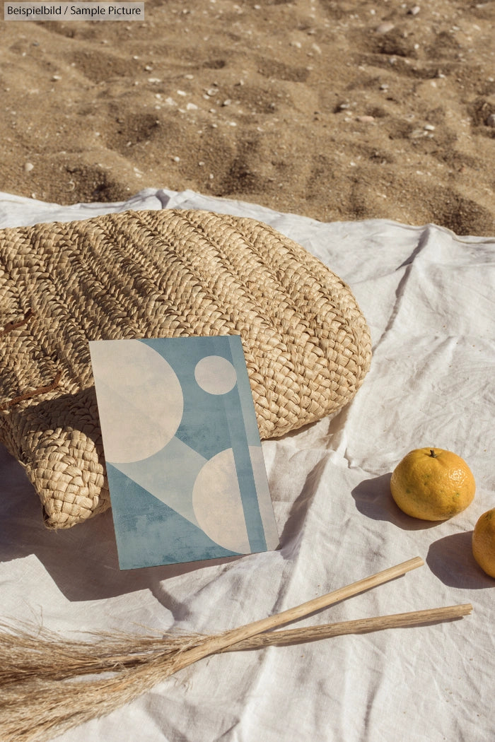 Woven bag and abstract art card on sandy beach with oranges and dried grass.