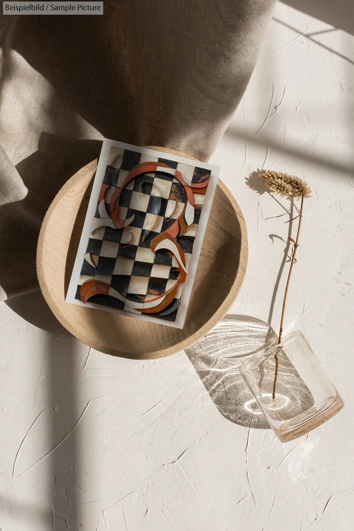 Abstract art print with curved patterns on a wood plate, beside dried flowers in a glass vase on a textured surface.