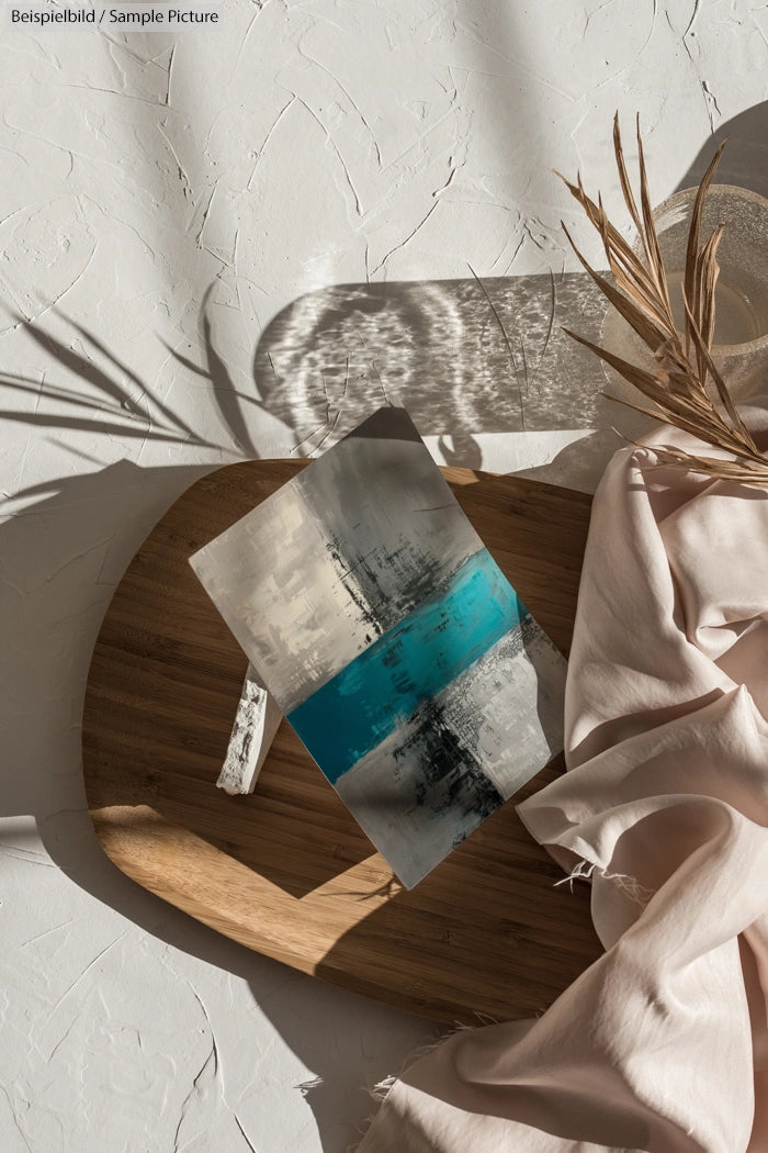 Abstract painting with gray, turquoise, and black colors on a wooden board, surrounded by cloth and dried plants.