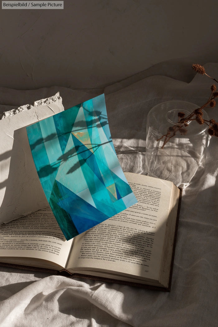 Geometric abstract artwork in blue and teal hues on textured paper placed on an open book in soft natural light.