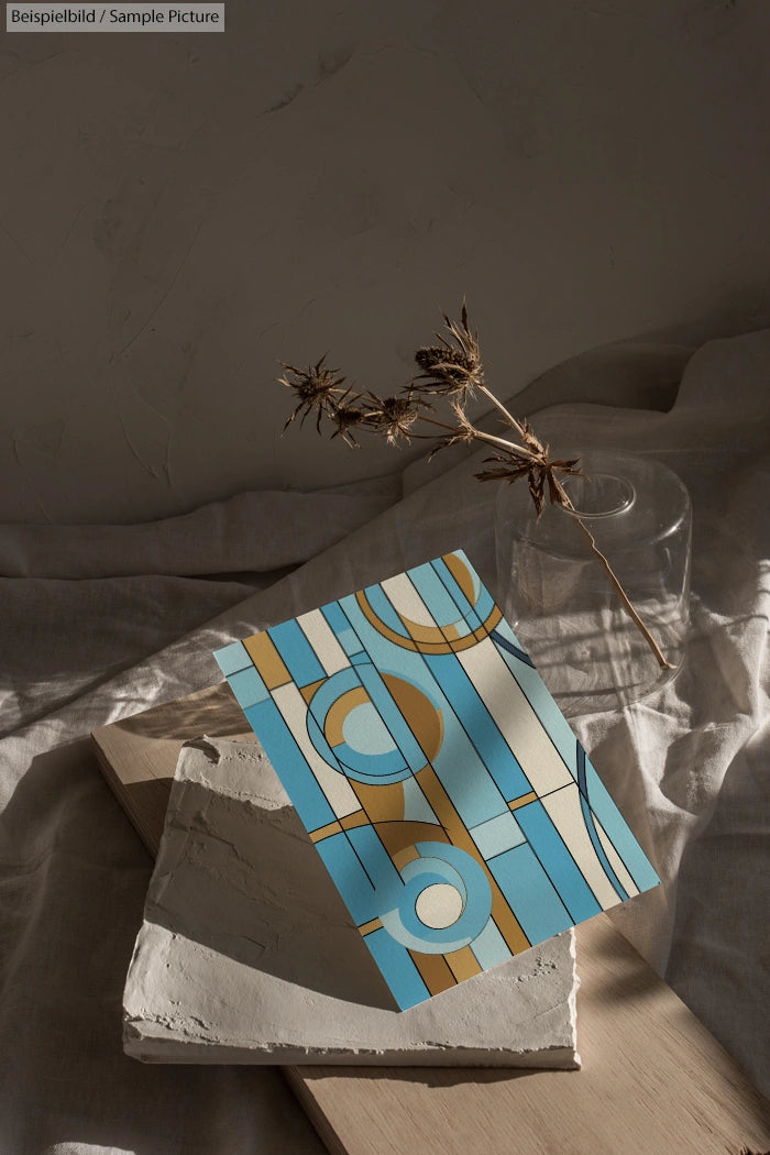 Abstract geometric artwork with circles and lines in blue and beige, displayed on textured fabric with dried plants.