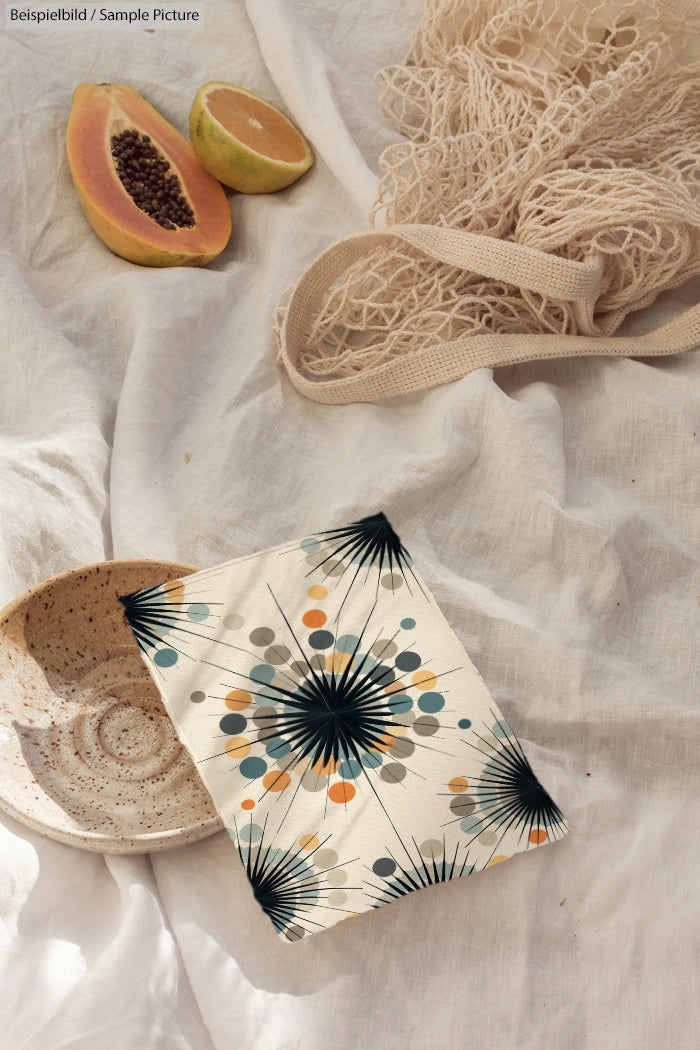 Fabric pouch with geometric floral print, papaya halves, net bag, and ceramic bowl on linen cloth background.