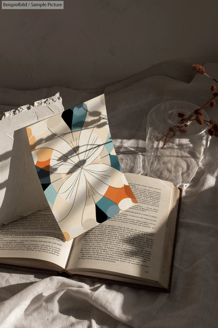 Open book on linen with abstract geometric print and dried flowers, natural light from window casting shadows.