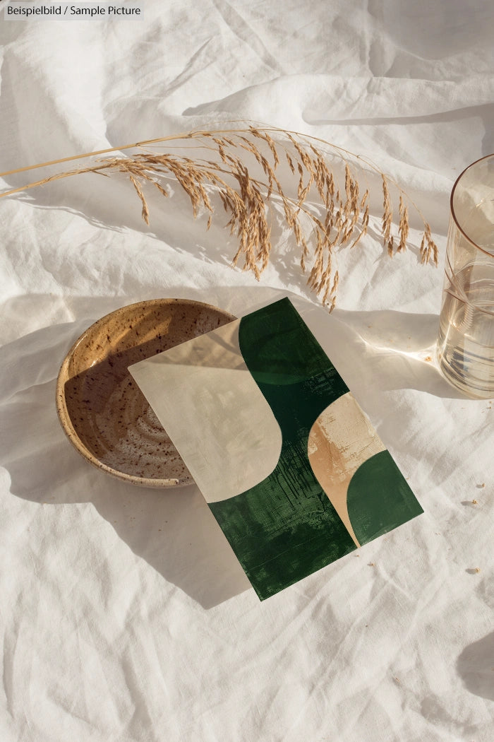 Abstract art card with green and beige shapes on soft fabric, next to a ceramic plate and a glass of water.