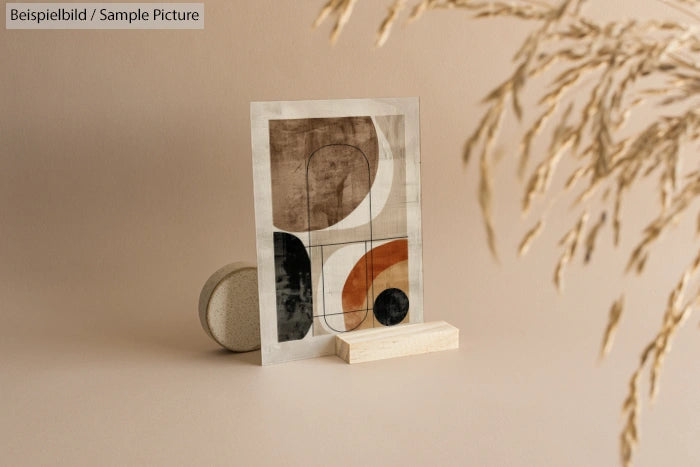 Abstract geometric art print with neutral tones on a minimal stand, surrounded by dried grass, on a beige background.