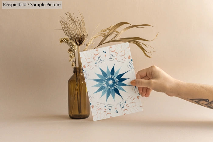 Hand holding art card with blue floral design near brown glass vase with dried plants.