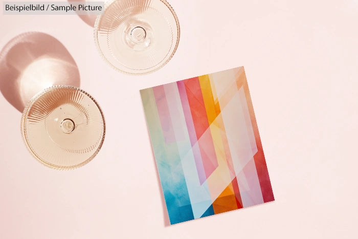 Colorful abstract geometric design on paper beside two empty champagne glasses on a soft pink surface.