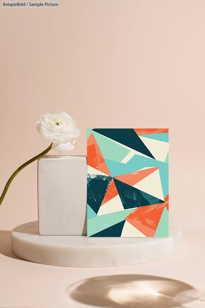 Abstract geometric art print with colorful triangles on a beige backdrop, next to a white flower in a simple vase.