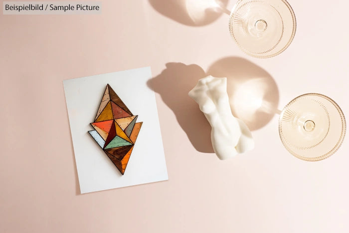 Geometric wood art piece on paper with a shadow of a sculpture and glass cups on a light pink surface.