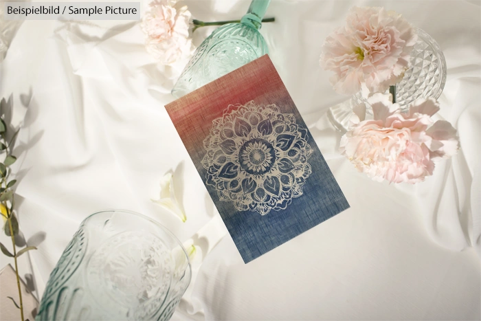 Intricate lace design card surrounded by pink carnations and glassware on white fabric backdrop.