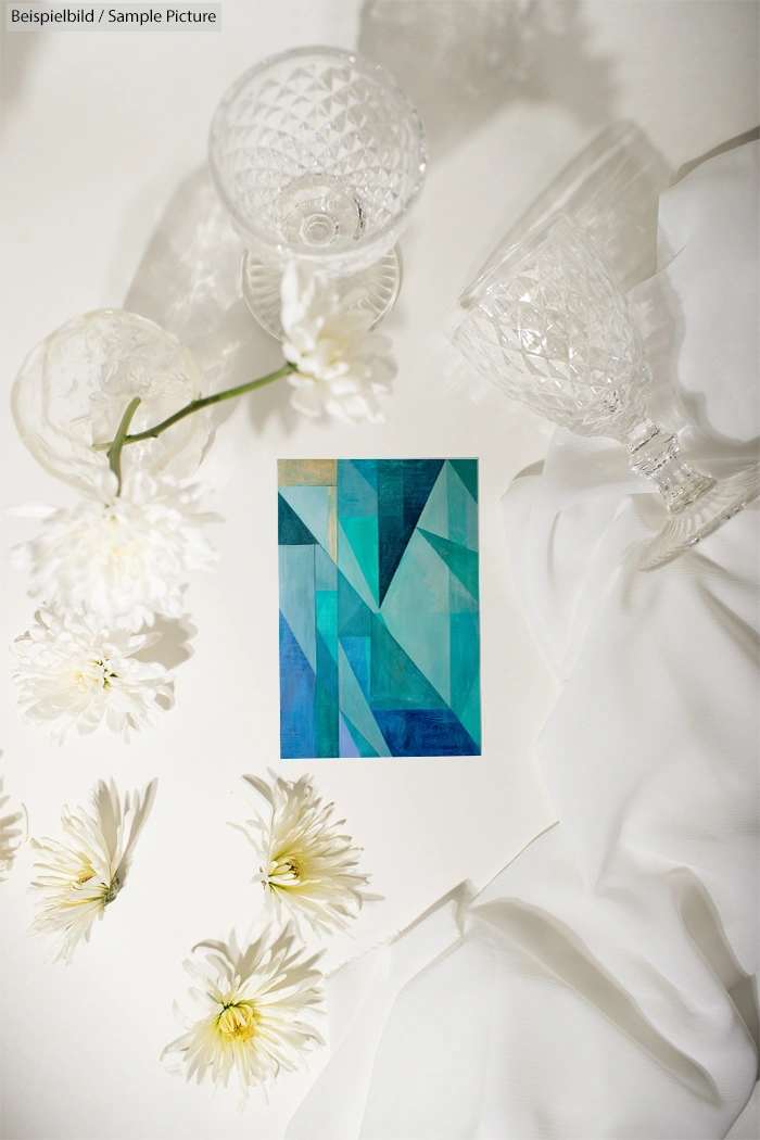 Geometric abstract painting with blue and green shapes, surrounded by crystal glassware and white flowers on a white cloth.